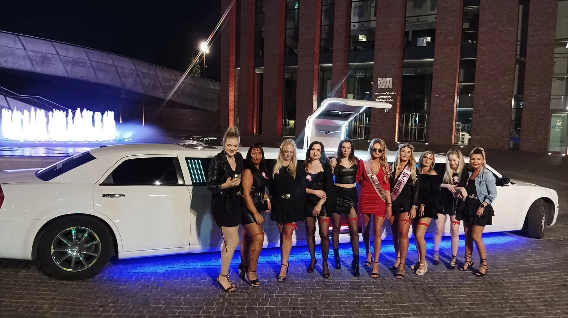 Grupa kobiet w wieczorowych strojach pozująca przy podświetlanej limuzynie na tle fontanny nocą.