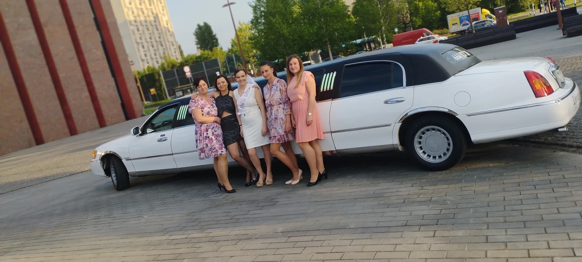 Pięć elegancko ubranych kobiet celebrujących wieczór panieński stojąc obok białej limuzyny.