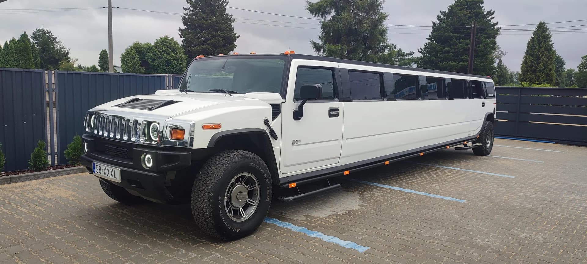 Biała limuzyna typu Hummer zaparkowana na wybrukowanym parkingu przy ogrodzeniu.