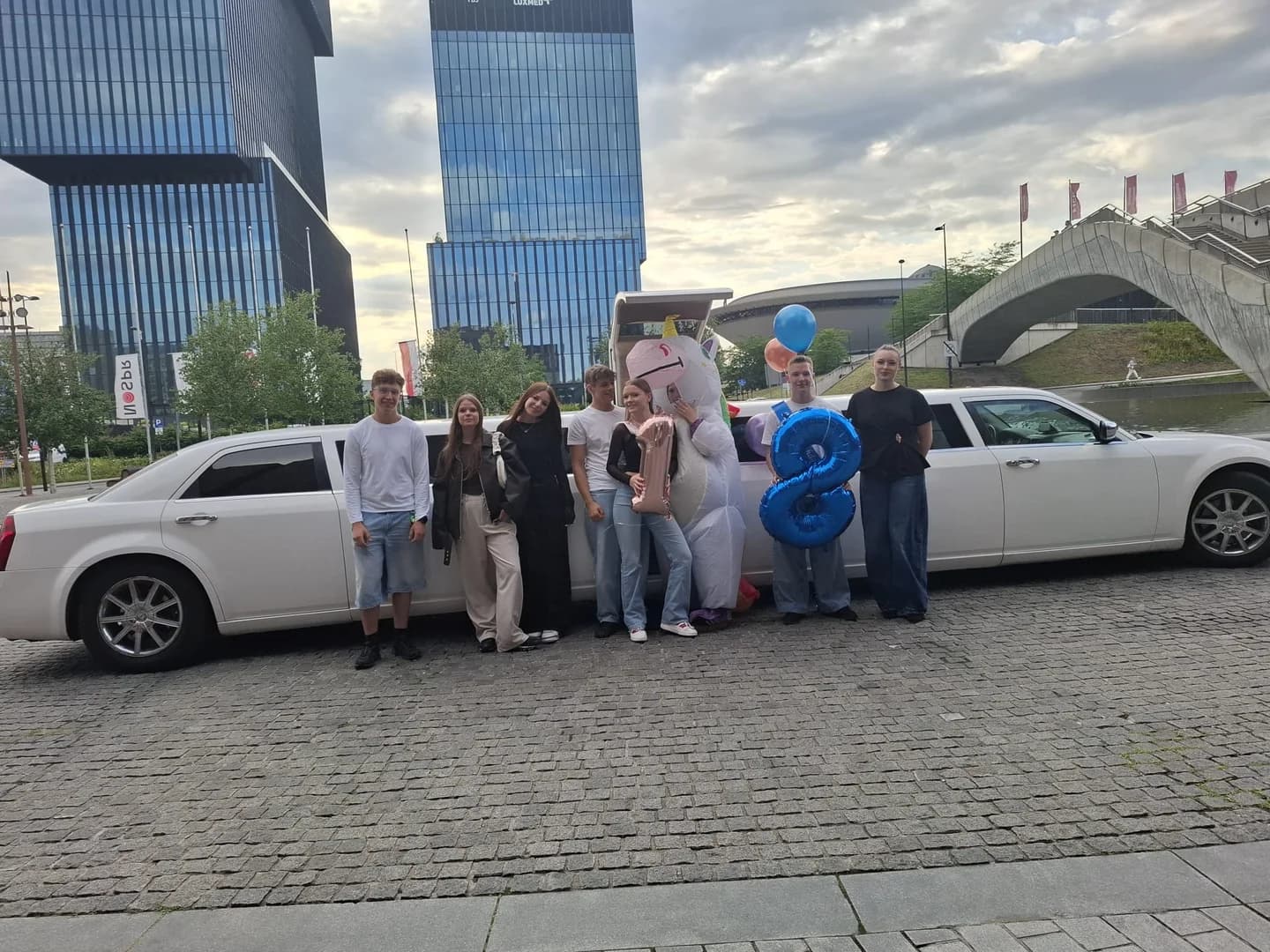 Grupa młodych osób, w tym jedna przebrana za jednorożca, pozująca z balonami w kształcie cyfry 18 przed białą limuzyną na tle nowoczesnych wieżowców.