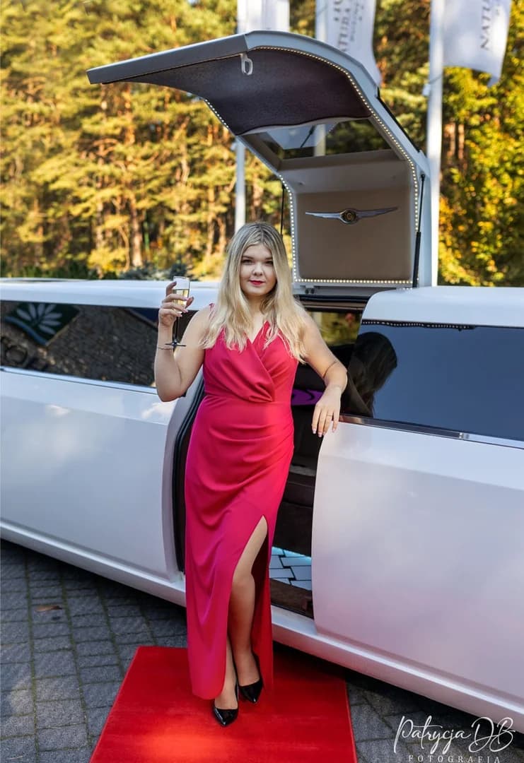 Blondynka w fuksjowej sukience stoi przy otwartej białej limuzynie, trzymając kieliszek szampana.