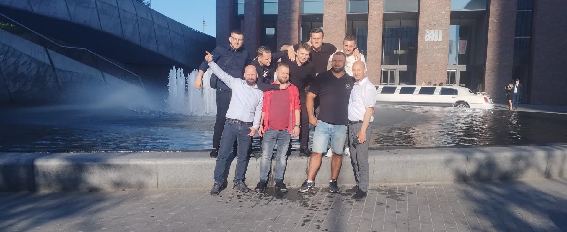 Grupa mężczyzn pozuje przy fontannie miejskiej, z białą limuzyną w tle.