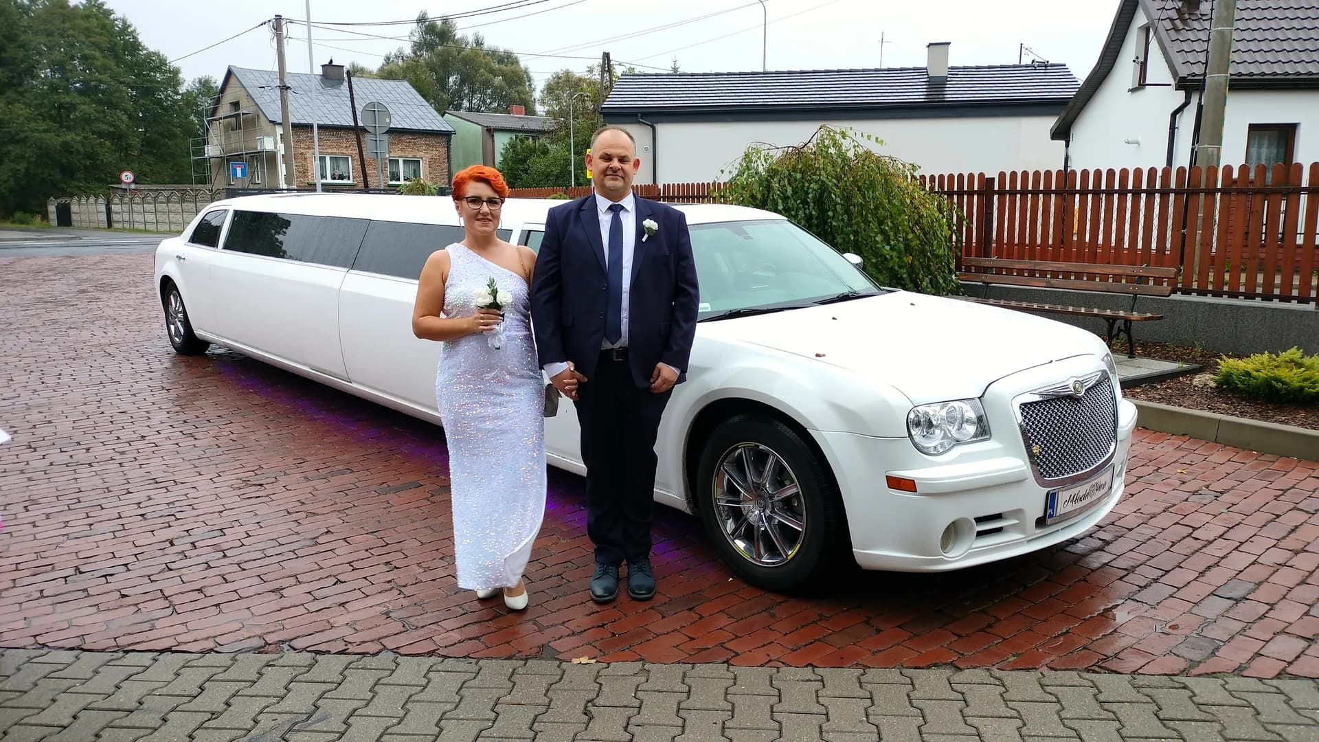 Para w eleganckich strojach pozująca przed białą limuzyną na brukowanym podjeździe.