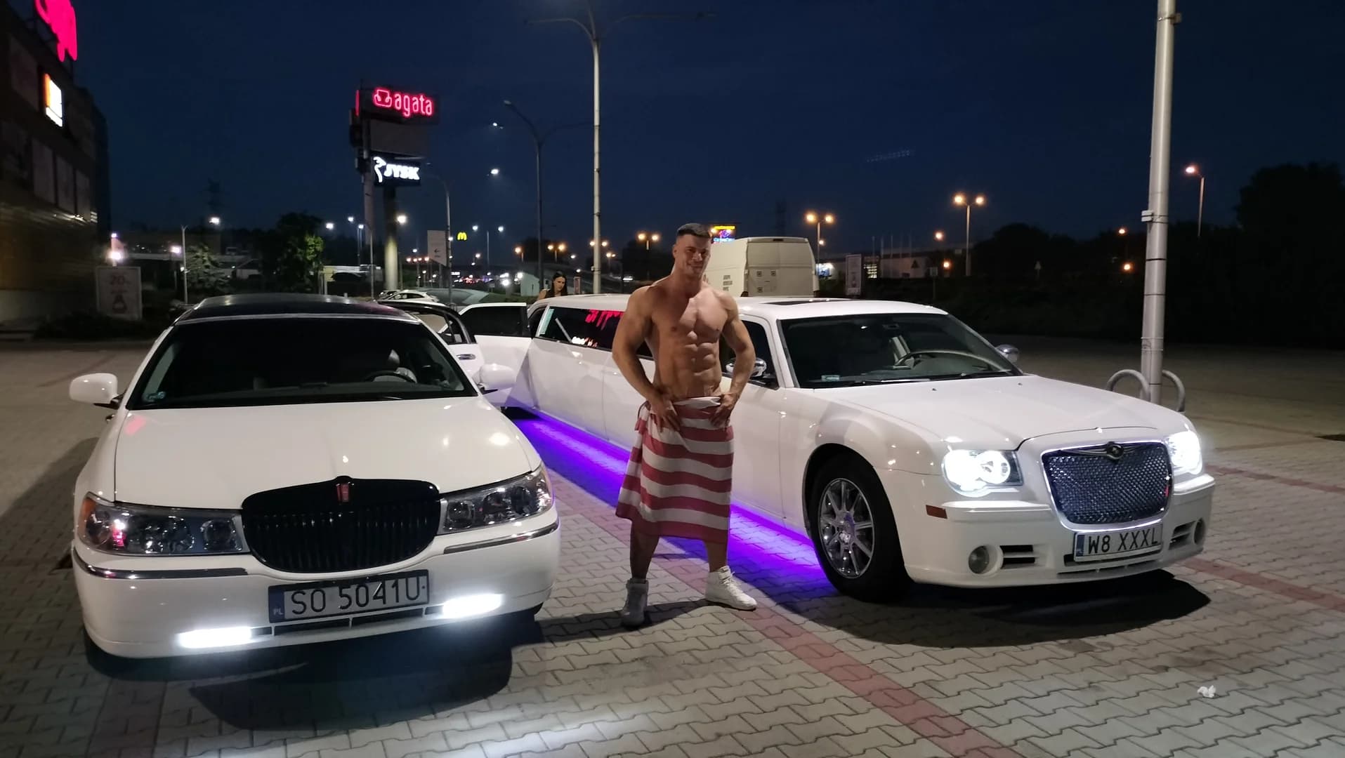 Mężczyzna bez koszulki pozuje nocą na parkingu między dwiema luksusowymi, białymi limuzynami.
