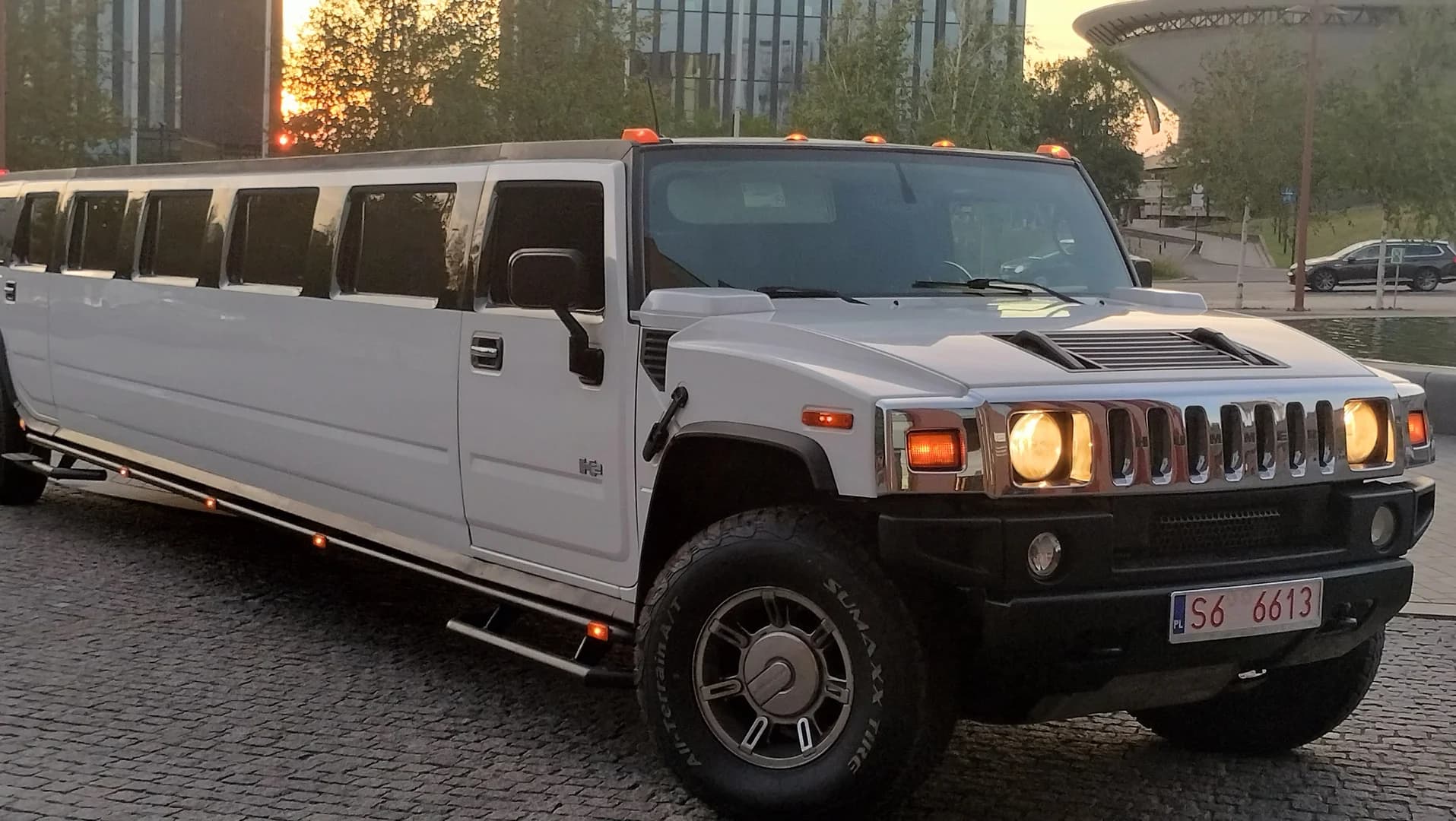 Biała limuzyna typu Hummer z zapalonymi światłami zaparkowana na brukowanej nawierzchni o zachodzie słońca.